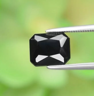 A faceted blue spinel gemstone held by tweezers against a green blurred background.